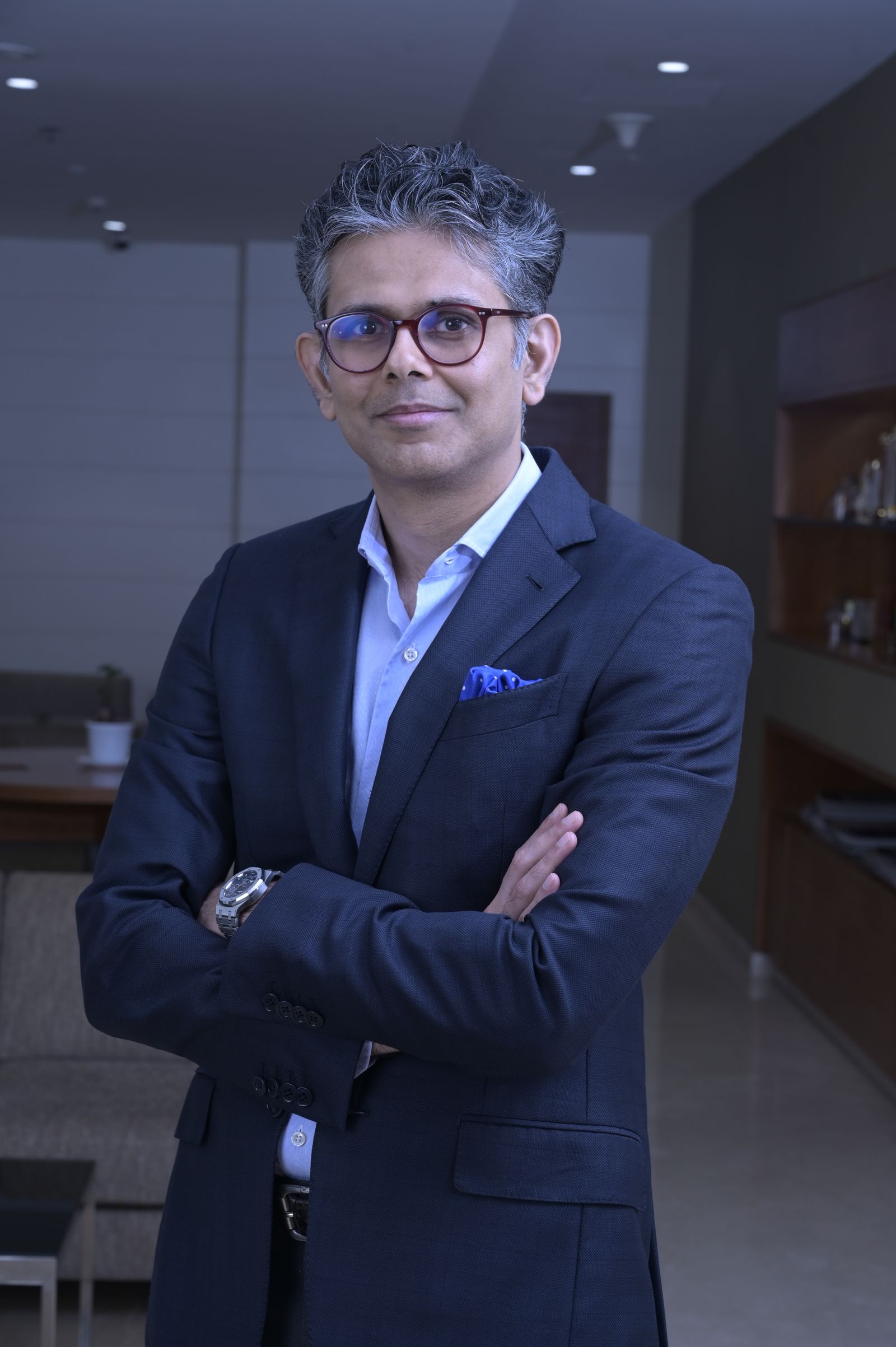 An executive in a dark suit stands with his arms folded in a corporate lobby and looks to the camera.